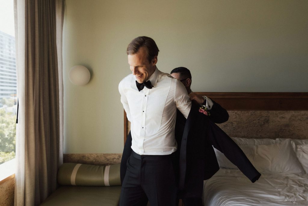 Groom putting on his black tuxedo jacket with the assistance of groomsman