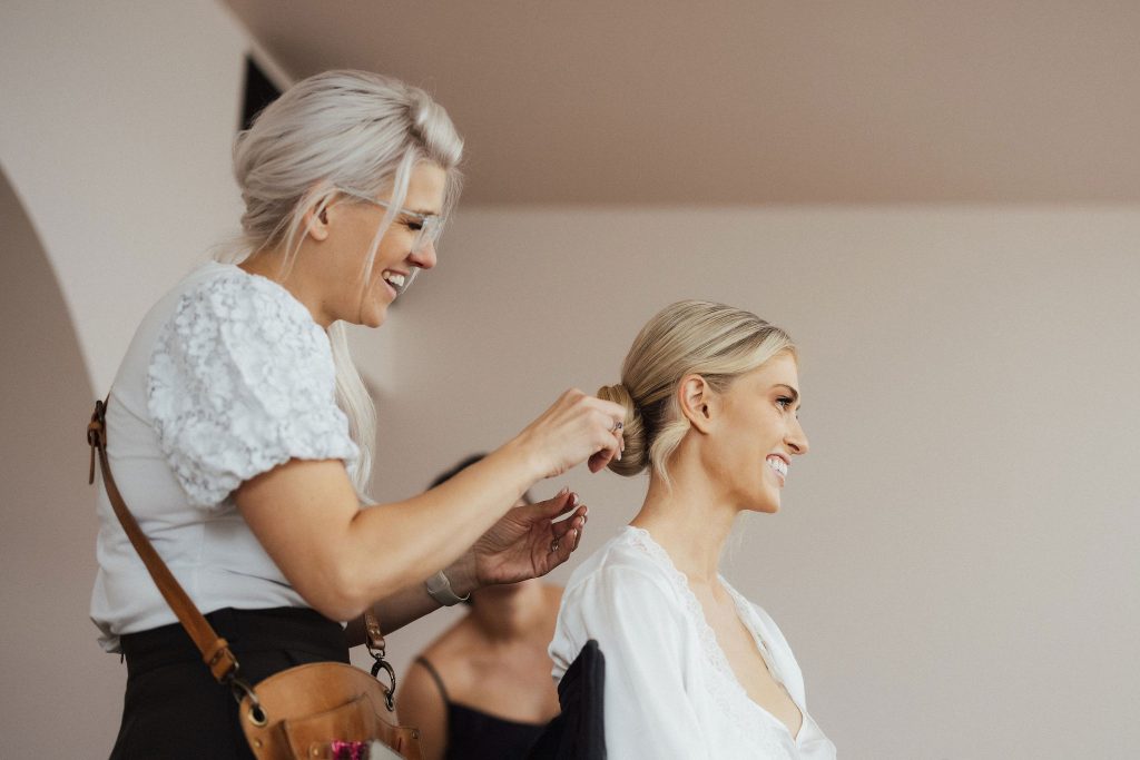 Bridal hair stylist putting final touches on blonde bride's low bun wedding hair style