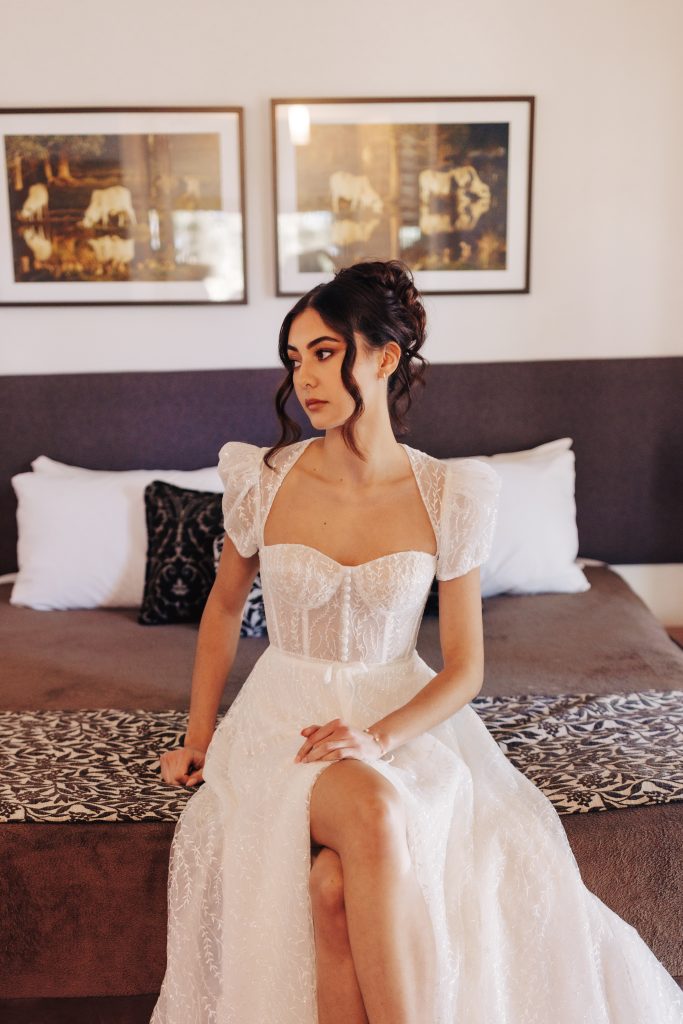 bride in pretty capped-sleeve bridal gown sitting on bed with wedding hair and makeup done in a luxury bridal suite cabin in Yandina Sunshine Coast